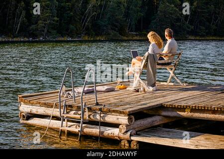 Man and woman using laptop while sitting on bench over pier against lake during summer Stock Photo