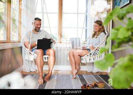 Smiling couple using laptops while sitting on rope swing in log cabin Stock Photo