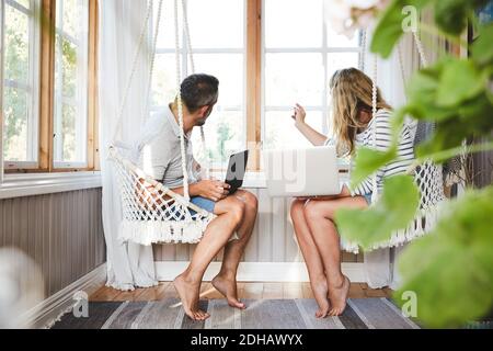 Man and woman looking through window while sitting with laptops in log cabin Stock Photo
