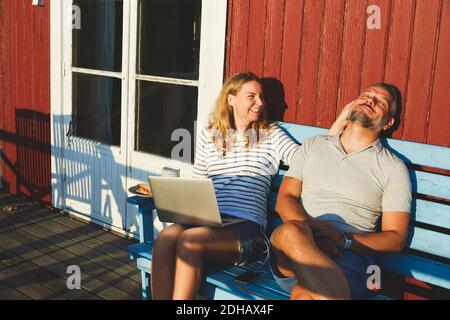 Happy woman using laptop while playing with man on bench at porch during summer Stock Photo