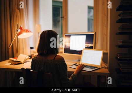 Rear view of girl using laptop and computer while sitting at illuminated desk Stock Photo