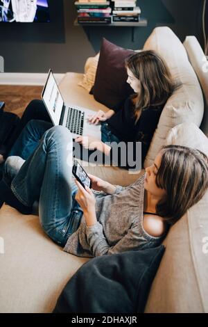 Smiling girls using laptop and mobile phone while resting on sofa at home Stock Photo