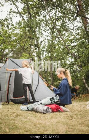 Teenage girl using phone while family pitching tent at camping site Stock Photo