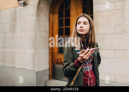 Thoughtful girl listening music through smart phone while walking by wall Stock Photo