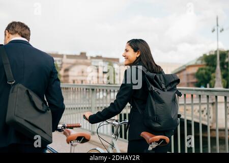 Smiling businesswoman looking at businessman while walking with bicycles on bridge in city Stock Photo