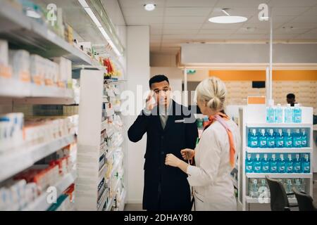 Male customer explaining headache to female pharmacist standing by rack at store Stock Photo