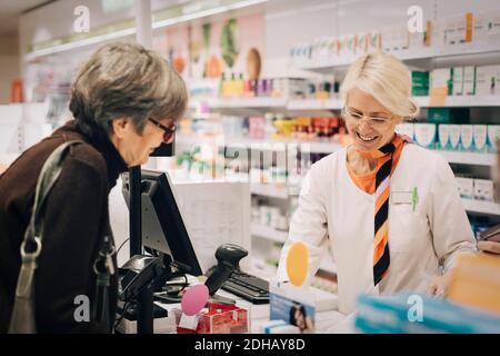 Smiling female owner standing with senior customer at checkout in pharmacy store Stock Photo