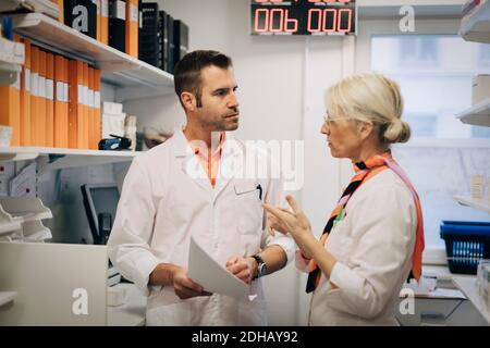 Mature medical colleagues discussing over documents by rack at pharmacy store Stock Photo