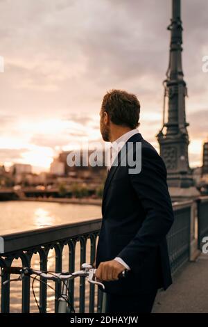 Side view of businessman with bicycle standing on bridge in city during sunset Stock Photo