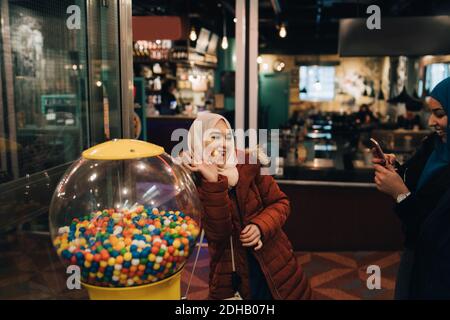 Young woman photographing cheerful female friend standing with bubble gum by vending machine Stock Photo