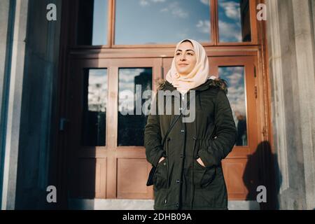 Confident young Muslim woman wearing hijab standing with hands in pockets against door Stock Photo