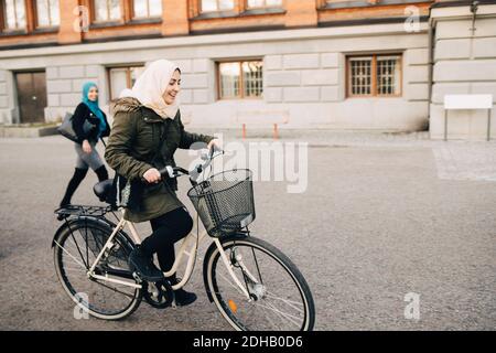 Happy young Muslim woman cycling on street with friend walking in city Stock Photo