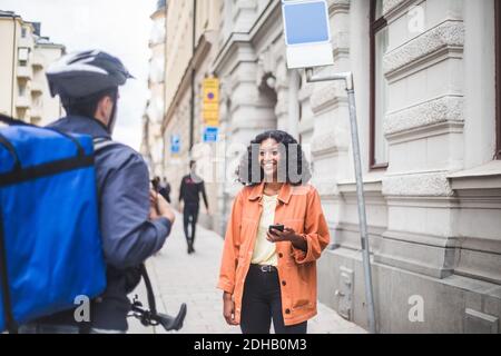 Smiling female customer talking with food delivery man while standing on sidewalk Stock Photo