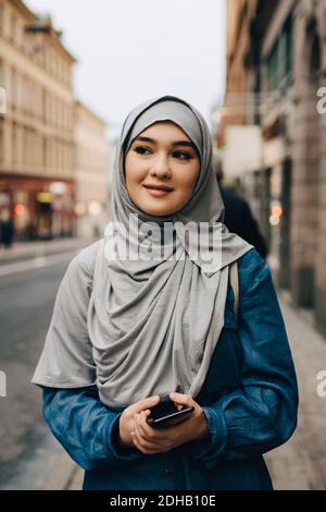 Young Muslim woman wearing hijab walking with mobile phone on sidewalk in city Stock Photo