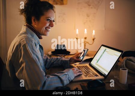Smiling engineer video conferencing on mobile phone while using laptop at desk in workshop Stock Photo