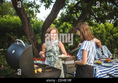 Smiling mother looking at daughter assisting in preparing food at barbecue grill in backyard during weekend party Stock Photo