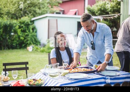 Woman looking at mature friend serving food on dining table in yard during weekend party Stock Photo