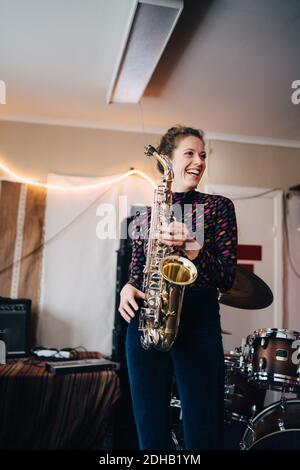 Happy young female musician playing saxophone in rehearsal studio Stock Photo