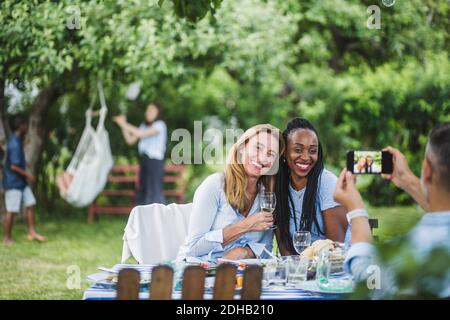 Man photographing friends with wine through mobile phone in garden party Stock Photo