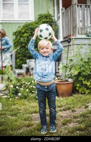 Full length of smiling boy holding soccer ball on head while standing in garden Stock Photo