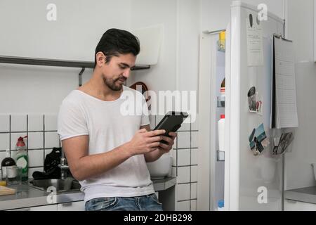 Young man using digital tablet while standing by open refrigerator at kitchen Stock Photo