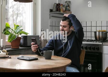 Man with headphones using mobile phone while sitting at table in kitchen Stock Photo