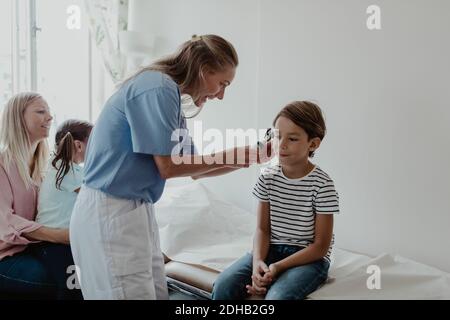 Female doctor examining boy's ear with otoscope while family sitting in medical room Stock Photo