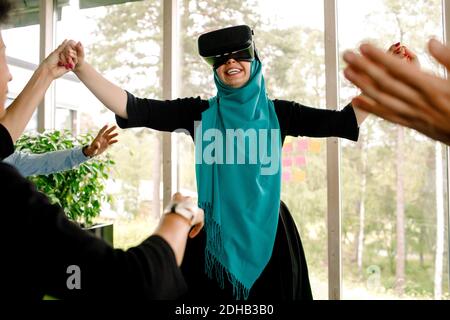Cheerful businesswoman holding hand of colleague while using virtual reality simulator during kick off meeting at conven Stock Photo