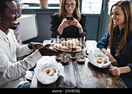 Smiling man giving buns to friend while woman using mobile phone at table Stock Photo