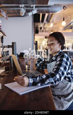 Smiling female manager talking on mobile phone while using laptop at bar counter Stock Photo