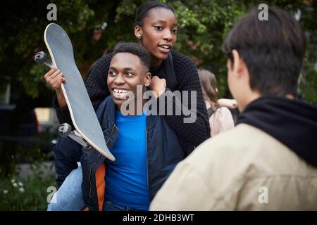 Smiling teenage boy piggybacking female friend holding skateboard Stock Photo