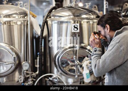 Mature man tasting from container while standing by storage tank in brewery Stock Photo
