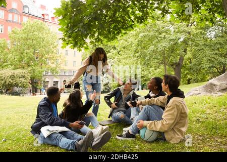 Happy teenage girl gesturing while standing by friends sitting on land at park Stock Photo