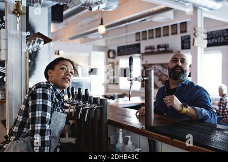 Female bartender with male customer looking up while standing at bar counter Stock Photo