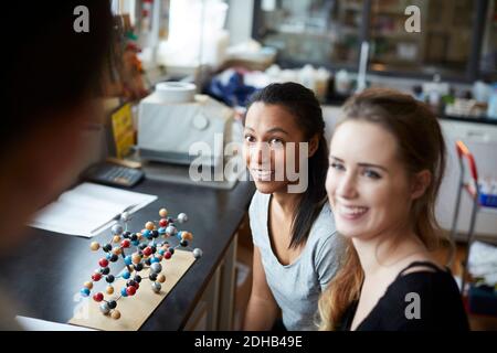 Smiling young female students by molecular structure looking at mature teacher in chemistry classroom Stock Photo