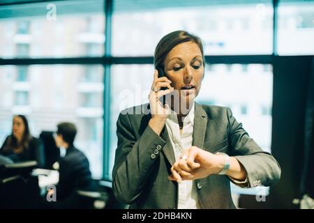Busy businesswoman talking on mobile phone while checking time on watch at office