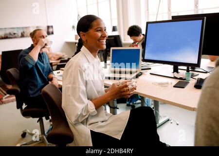 Smiling businesswoman having drink looking away while sitting with colleague at office desk Stock Photo
