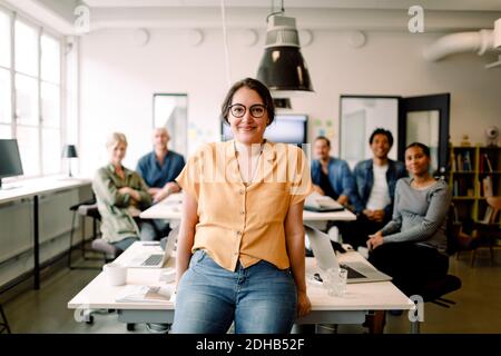 Portrait of smiling female professional leaning on table against executives in modern office Stock Photo
