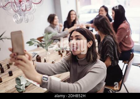Smiling young woman taking selfie with female colleagues sitting at table in perfume workshop Stock Photo