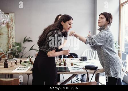 Young woman smelling perfume fragrance from female colleague's wrist while standing at workshop Stock Photo