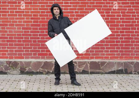 A young man stands against a background of a red brick wall and holds in front of him two white blank canvases for painting Stock Photo