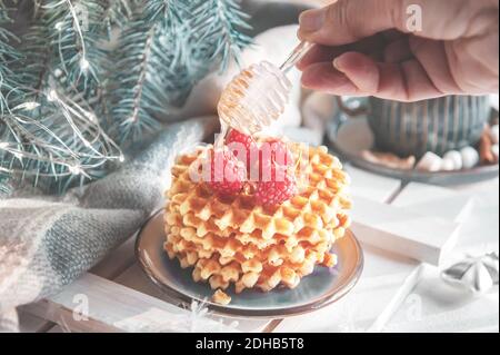 Viennese waffles with raspberries and honey on the Christmas table with a mug of cocoa and marshmallows in the background. The hand is pouring honey o Stock Photo