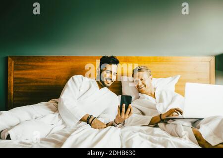Smiling man showing smart phone to boyfriend using laptop while leaning in bed at hotel Stock Photo
