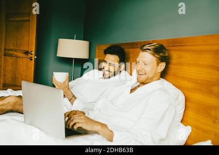 Smiling gay couple using laptop while leaning on bed in hotel room Stock Photo