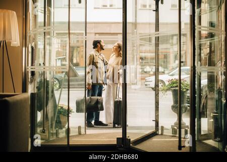 Couple with luggage talking while standing at doorway of hotel seen through glass Stock Photo