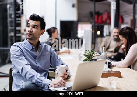 Thoughtful businessman having coffee and using laptop while sitting with colleagues at table Stock Photo
