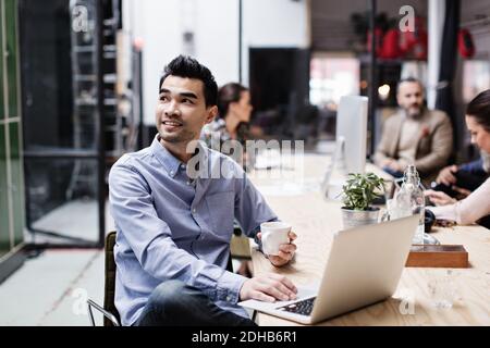 Smiling businessman having coffee and using laptop while sitting with colleagues at table Stock Photo