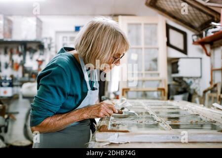 Side view of senior female owner scraping window frame on workbench at store workshop Stock Photo