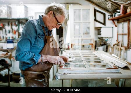 Side view of senior male owner scraping window frame on workbench at store workshop Stock Photo