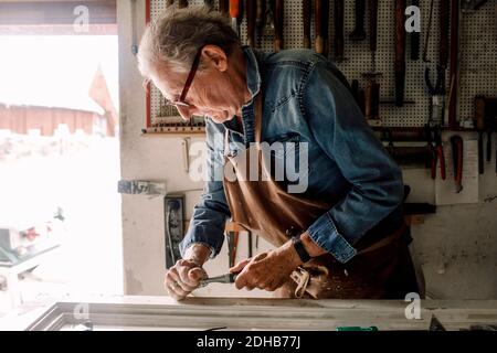 Senior male owner scraping window frame on workbench at store workshop Stock Photo